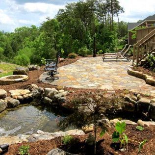 A backyard patio with flagstone paving, a small pond, a fire pit, and lounge chairs surrounded by trees.