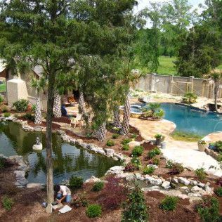 A person gardens near a pond and swimming pool in a landscaped backyard with stone patios and trees.