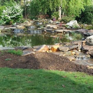 A tiered rock garden pond with a small waterfall, surrounded by dark mulch, green grass, and lush trees.
