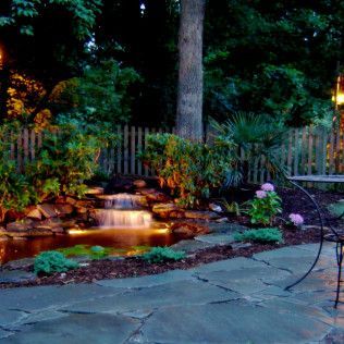 A landscaped backyard at night featuring a stone patio, a small glowing waterfall pond, lush greenery, and a wooden fence.