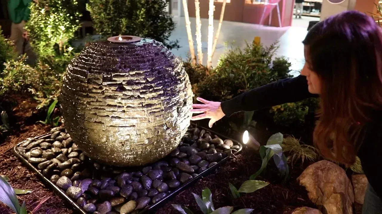 A person reaches out to touch a textured, wet spherical water fountain surrounded by dark pebbles in a garden setting.