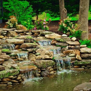 A multi-tiered stone waterfall feature cascading into a small pond, surrounded by lush green foliage and trees.