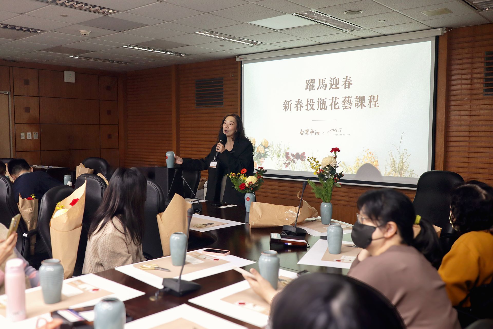一位女士在教室研討會上發言，聽眾坐在課桌前，面前是投影演示螢幕。