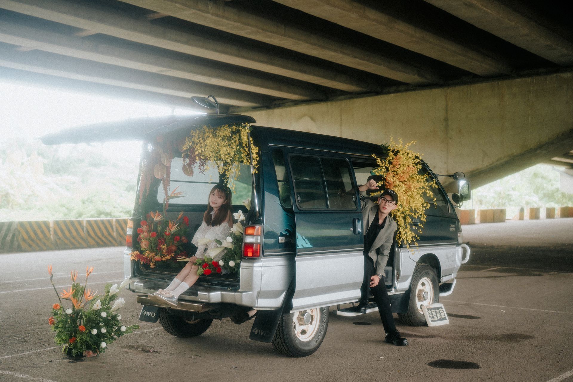 橋下停著一輛裝飾著鮮花的銀色廂型車，車旁坐著兩個人，旁邊站著一個人。