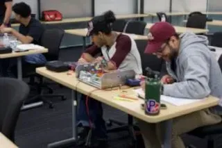 Students working at desks in a classroom, focused on laptops and notebooks.