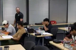 Classroom with several students seated at desks while a teacher stands at the back.
