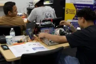 People seated at a table in a workshop, working with tools and electronics prototypes.
