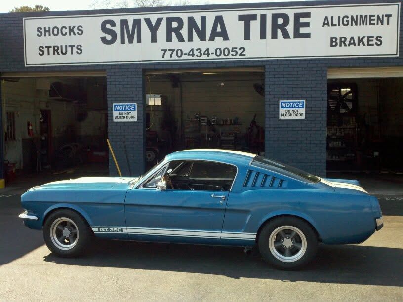 Blue classic car parked outside Smyrna Tire shop with service signs for shocks, struts, alignment, and brakes