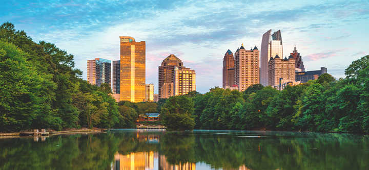 City skyline glowing at sunset above a calm lake, framed by lush green trees.