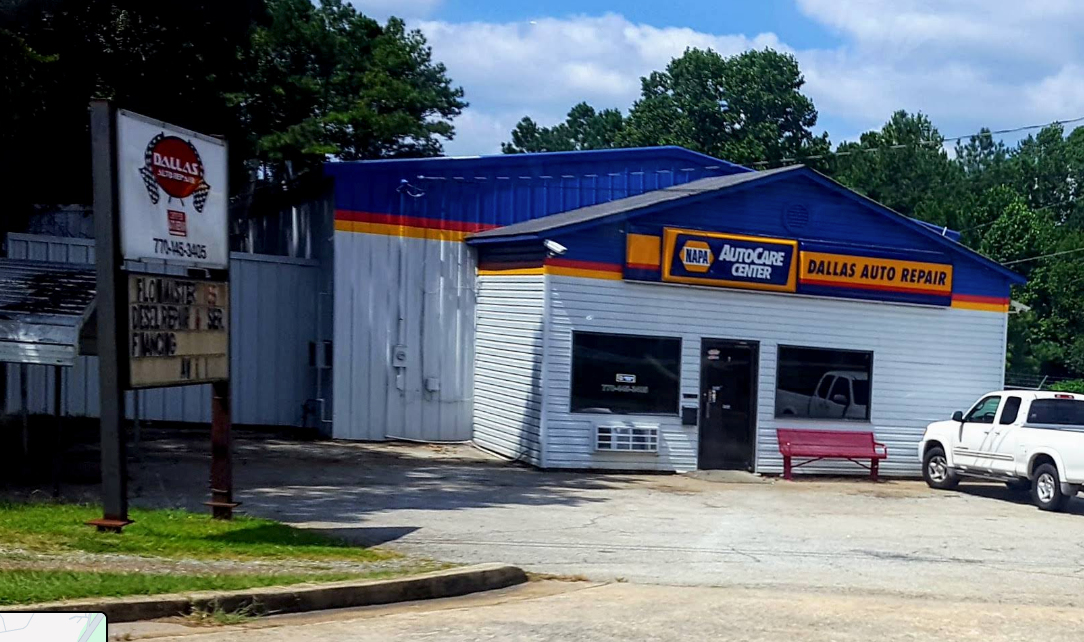 Small auto repair shop with blue roof, white walls, and parked vehicles on a sunny street corner.