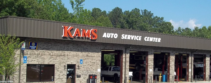 KAMS Auto Service Center storefront with stone facade and red sign under a wooded hillside