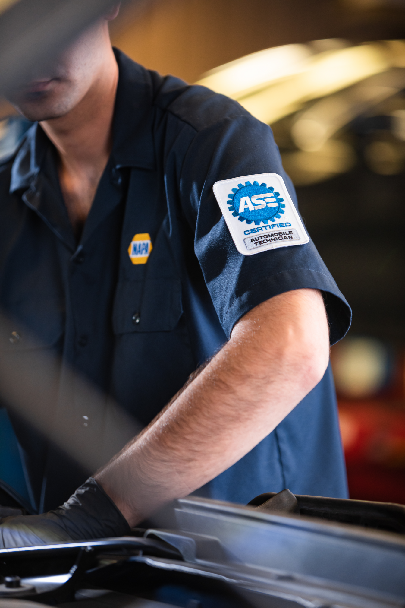 Mechanic in dark uniform working under a car hood with an ASE patch on the sleeve