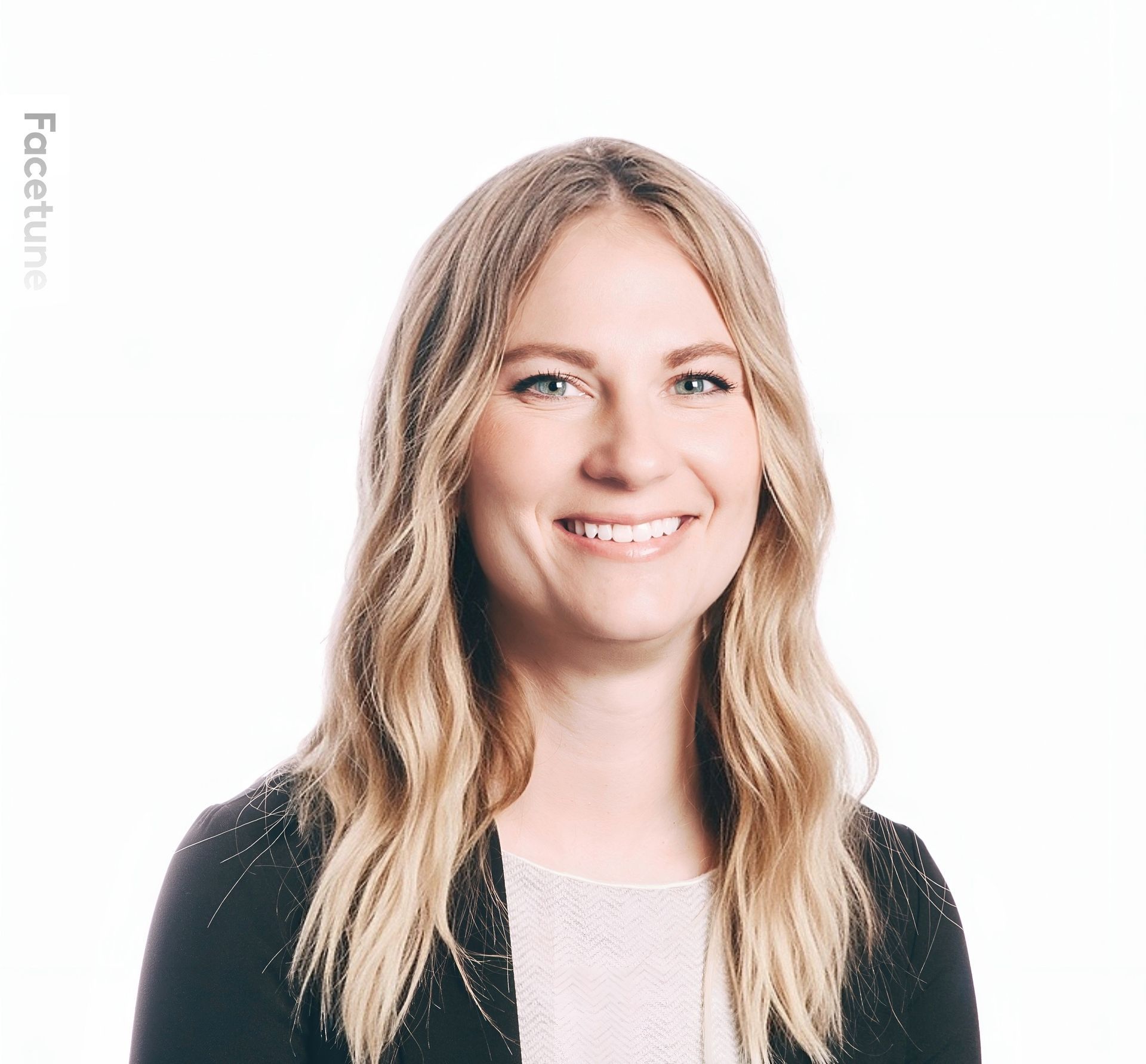 A smiling person with long blonde hair, wearing a black blazer over a white shirt against a plain white background.