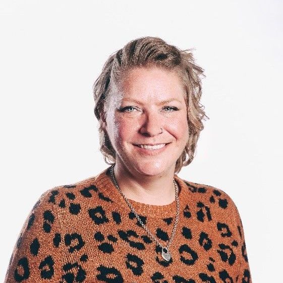 A smiling person with wavy hair wearing a brown leopard-print sweater and a silver chain necklace against a white background.