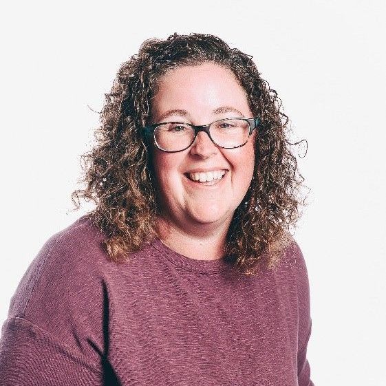 Woman with curly brown hair and glasses smiles, wearing a purple shirt.