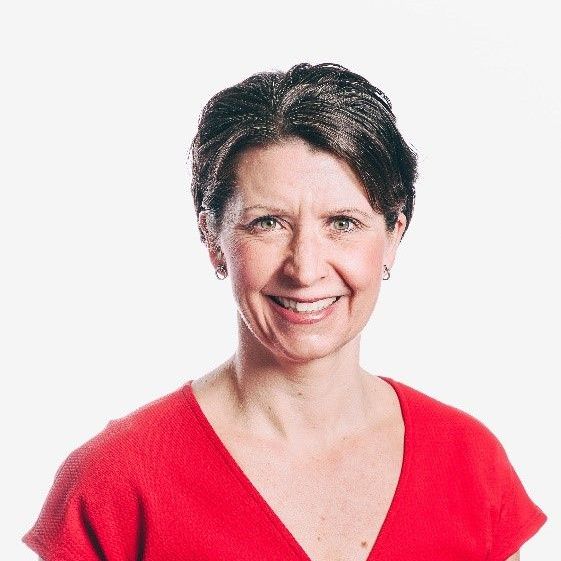 A person with short brown hair smiling while wearing a red V-neck shirt, set against a plain white background.