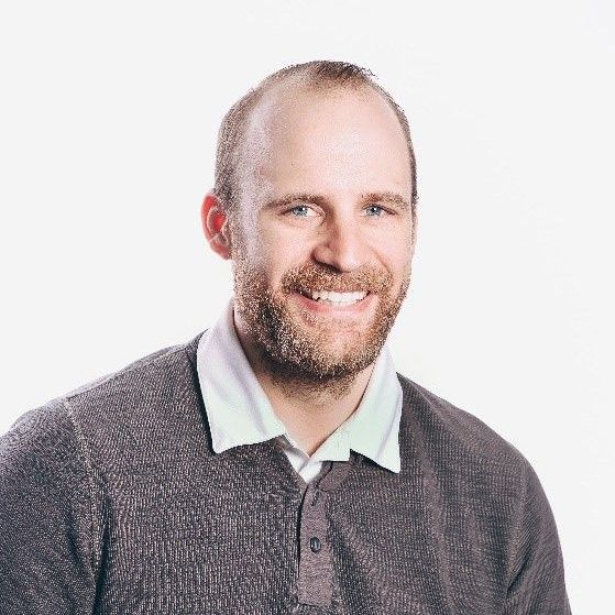 Man with a beard smiling, wearing a grey sweater over a white collared shirt, against a white background.