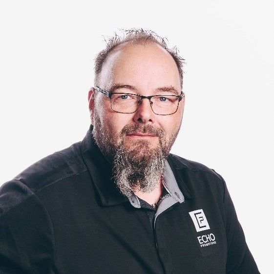 Man with beard and glasses in black polo shirt with logo.