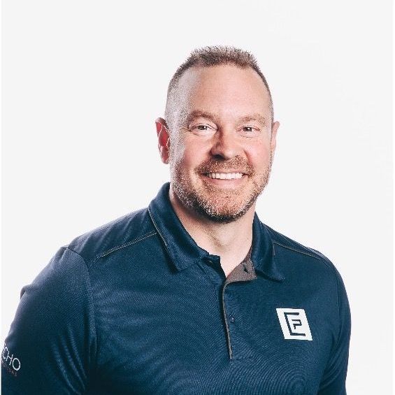 A person with a beard smiling against a plain white background, wearing a dark blue polo shirt with a logo on the chest.
