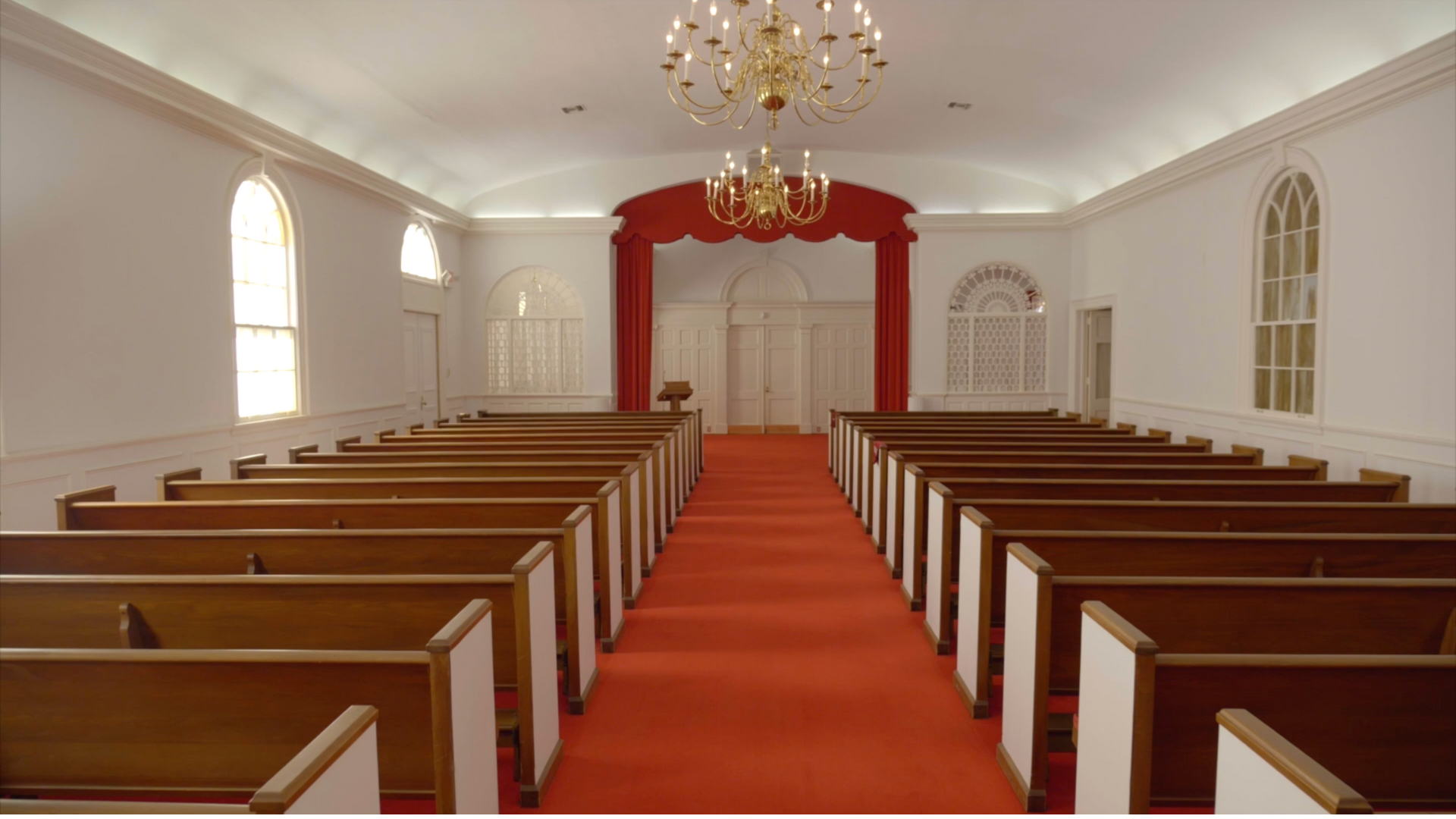 Inside a church. Rows of wooden pews face a red carpet leading to a red curtain and doors. Bright.