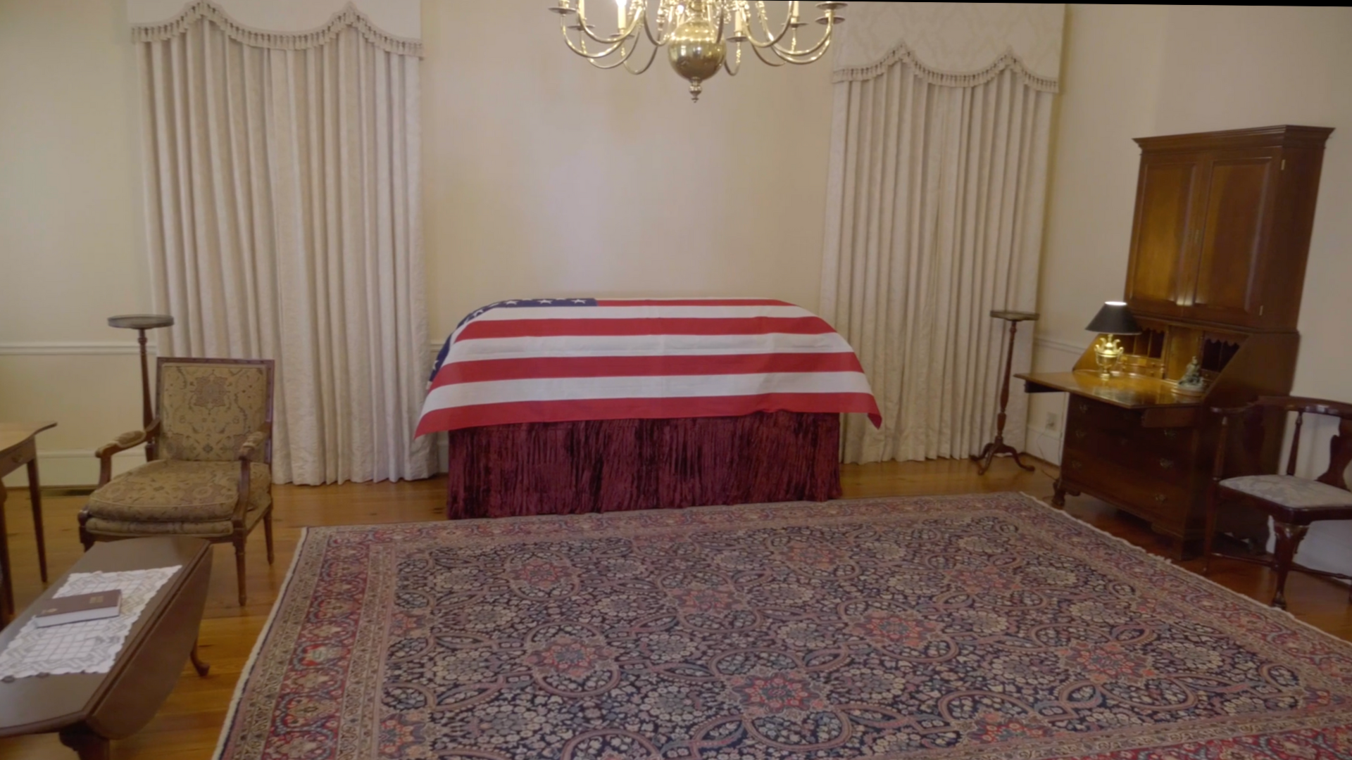 A casket draped with the American flag in a room with curtains and an oriental rug.