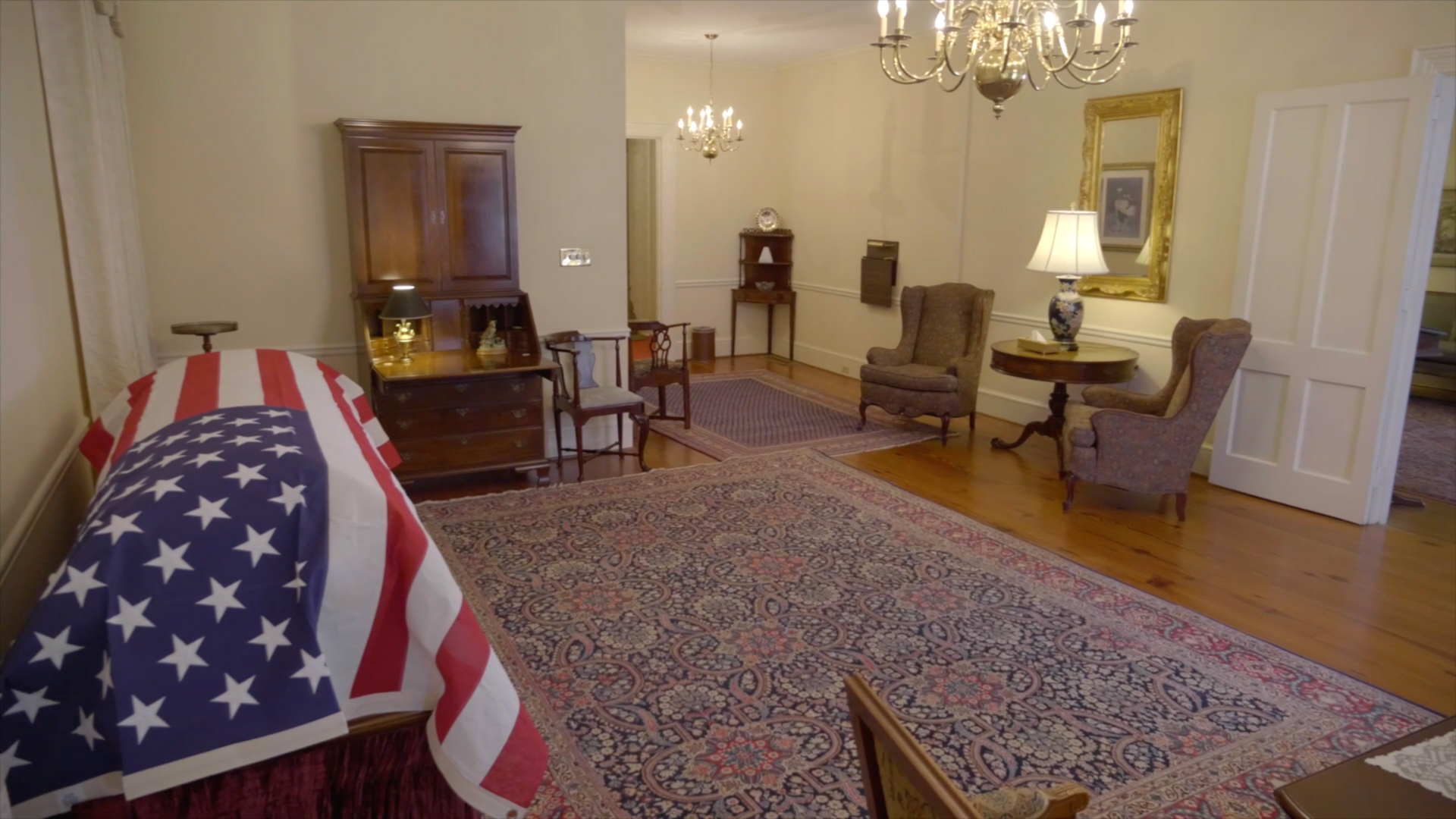 A room with a casket draped in an American flag, antique furniture, and a chandelier.