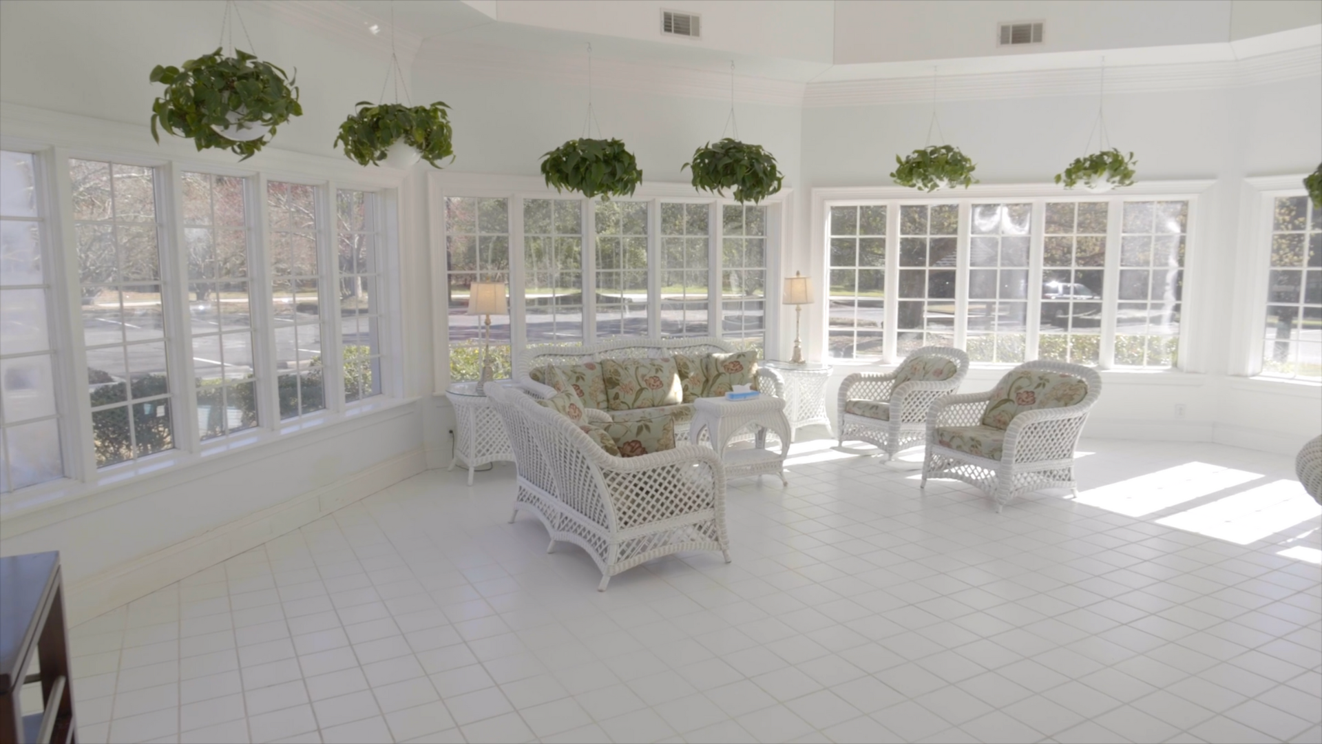 White sunroom with wicker furniture, hanging plants, and large windows.