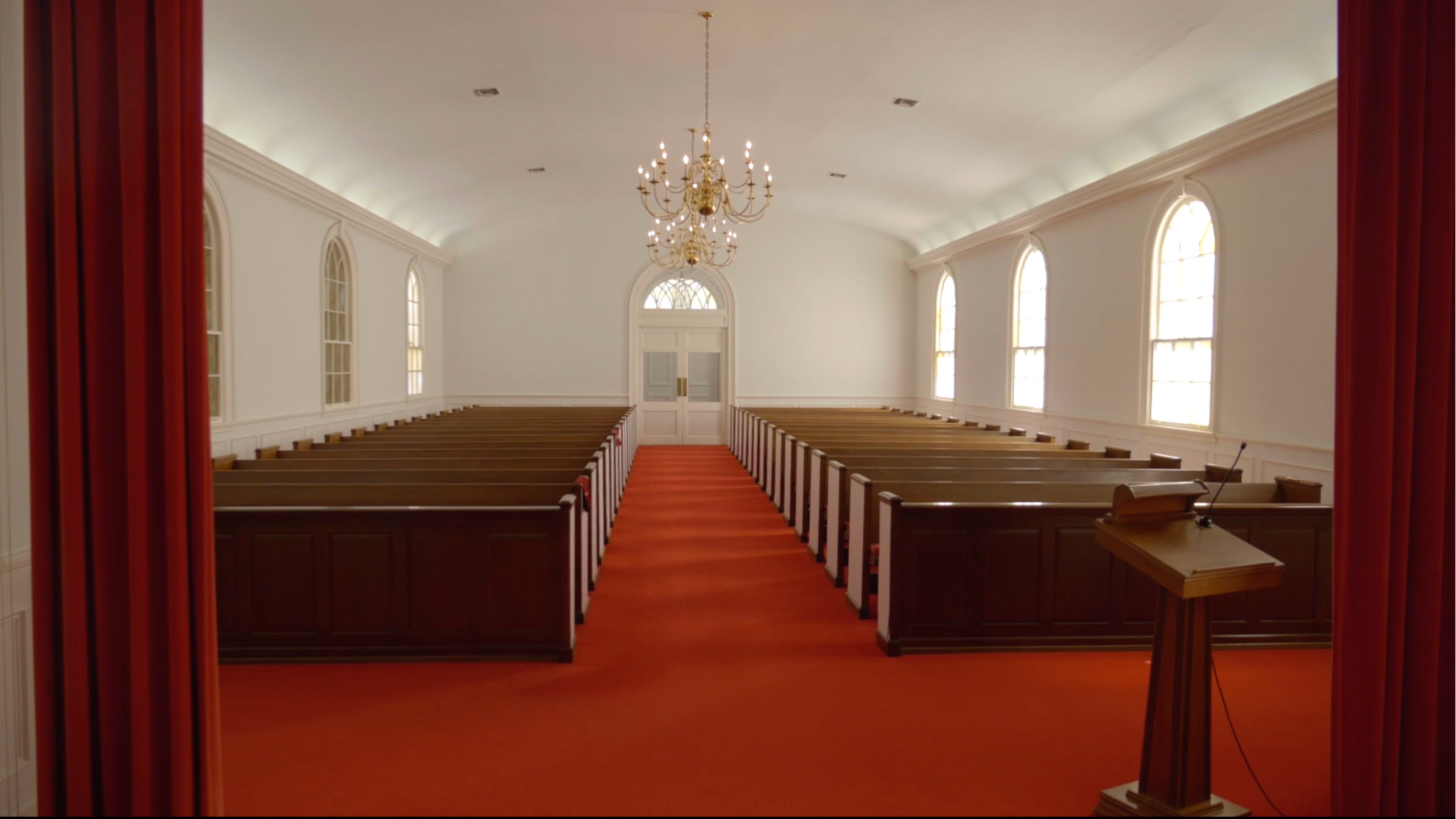 Interior of a church with rows of wooden pews, red carpet, and a pulpit.  Light from windows and a chandelier.