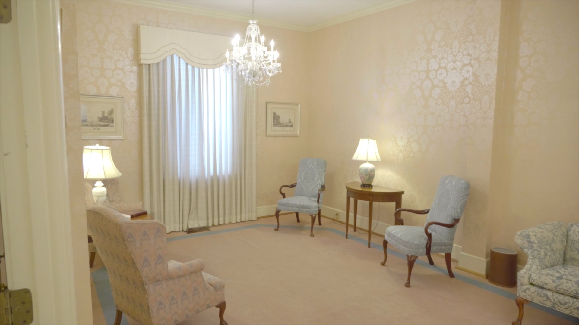 A light pink room with ornate wallpaper and furniture, chandelier, and window with drapes.