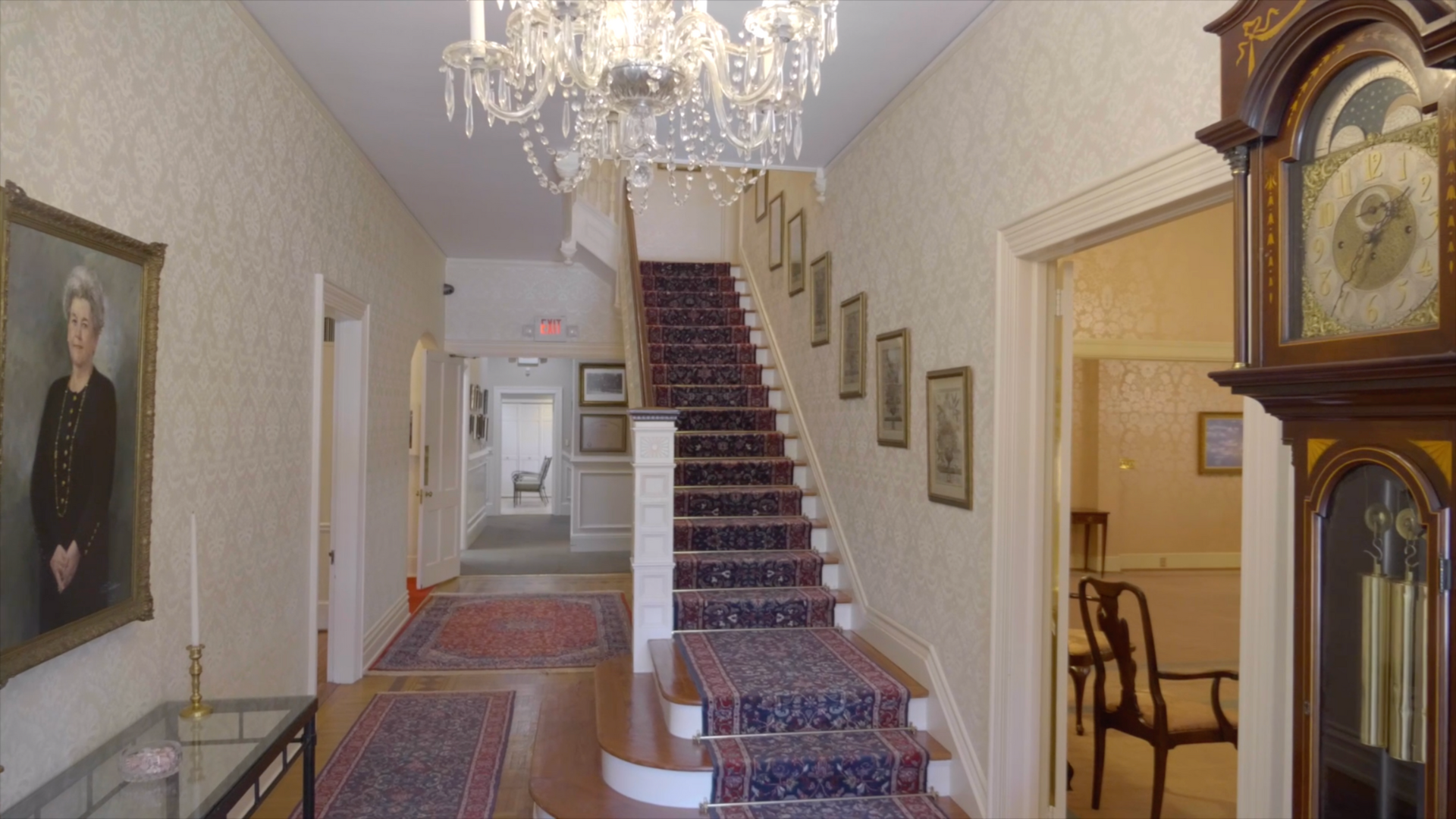 Elegant hallway with staircase, chandelier, grandfather clock, and portrait.