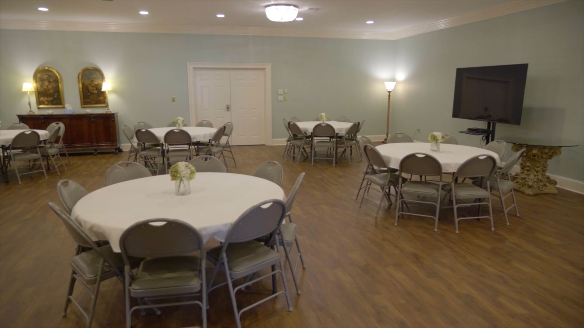 A large room with round tables, chairs, a TV, and artwork. Light blue walls, wood floors, and neutral colors.
