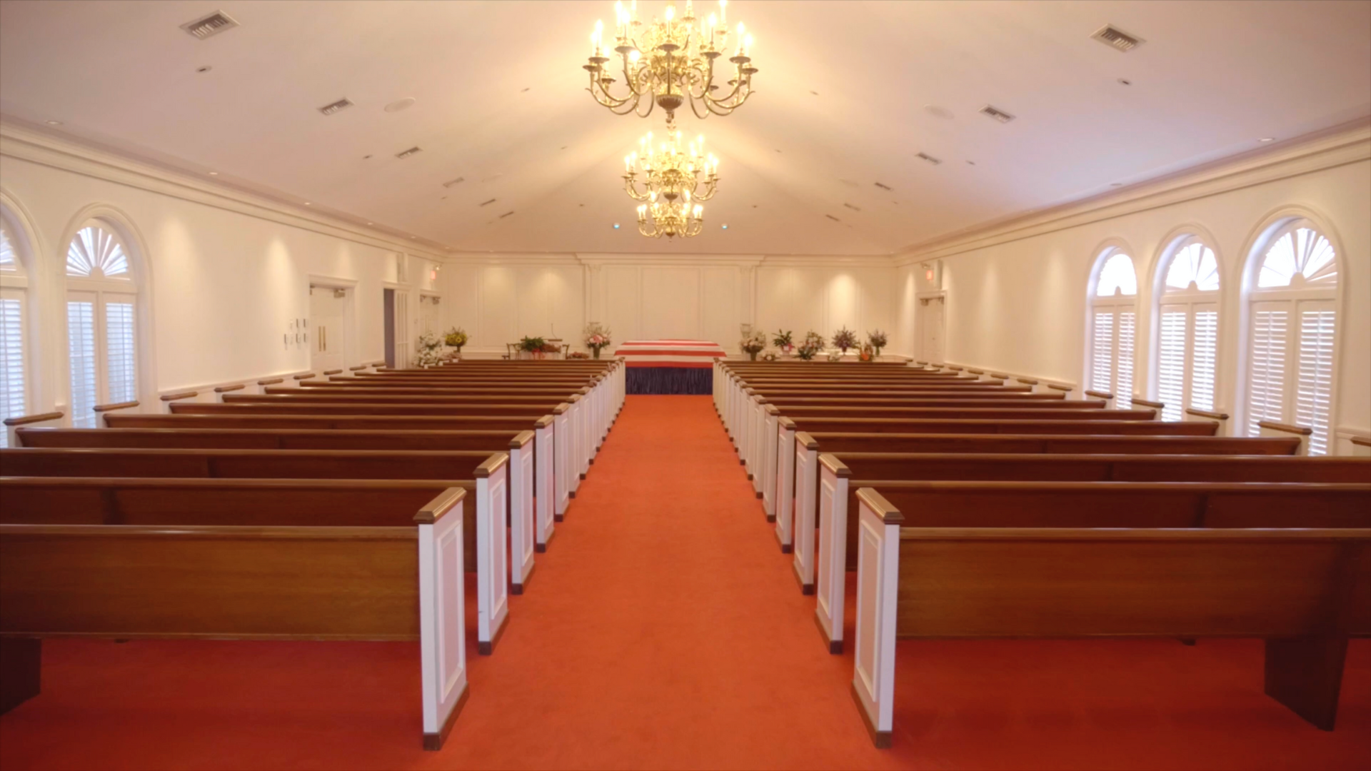 Interior of a bright chapel with rows of wooden pews, an altar, and two chandeliers.