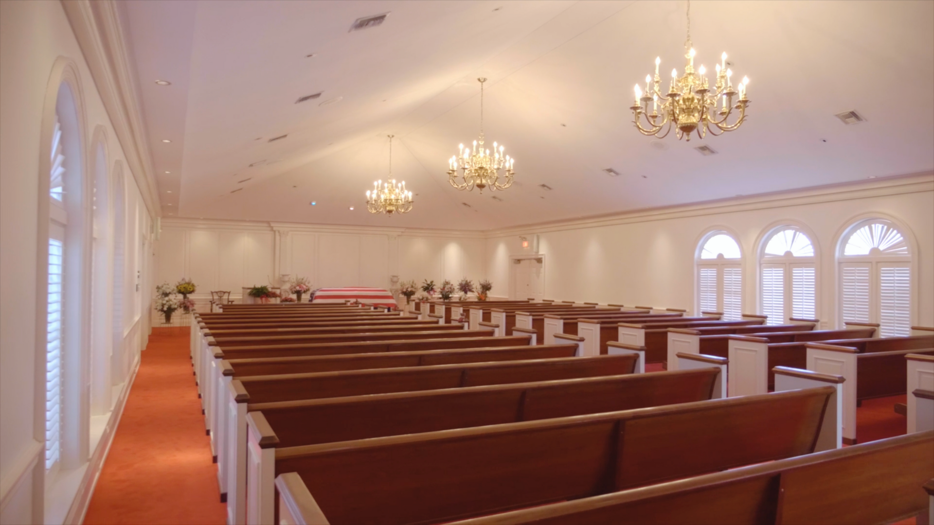Empty church interior with rows of wooden pews, three chandeliers, and arched windows.