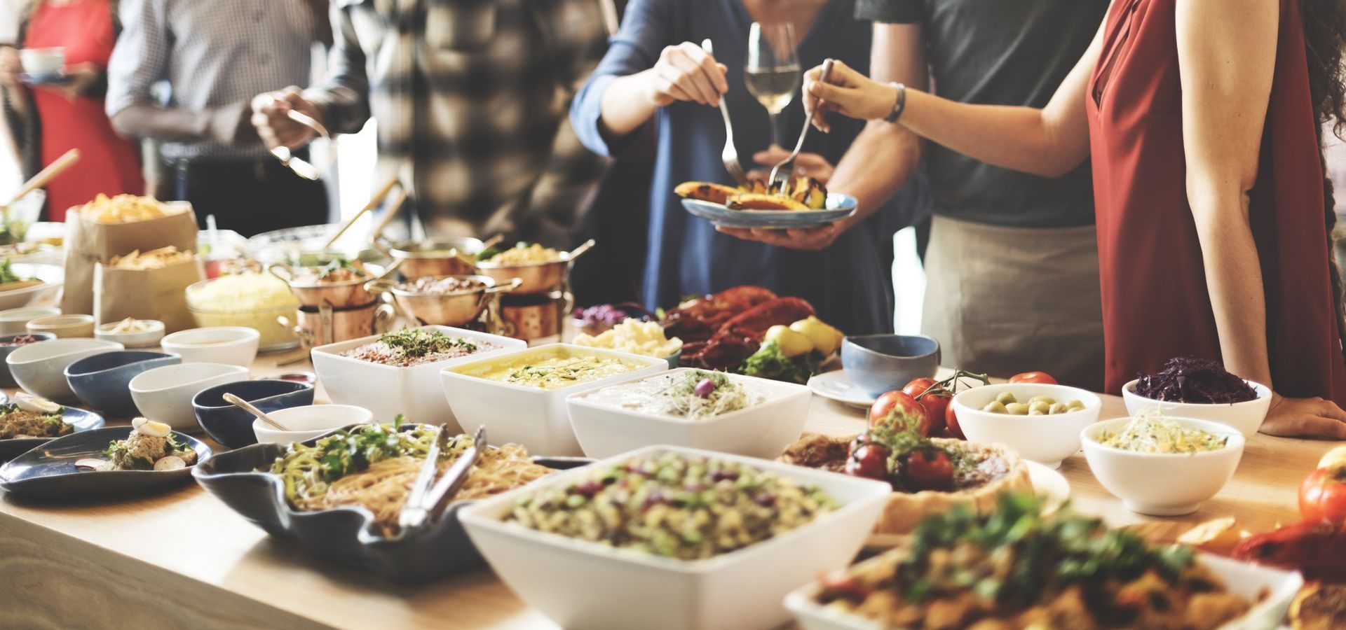 People at a buffet table, selecting food. Various dishes in white and black bowls on a wooden surface.