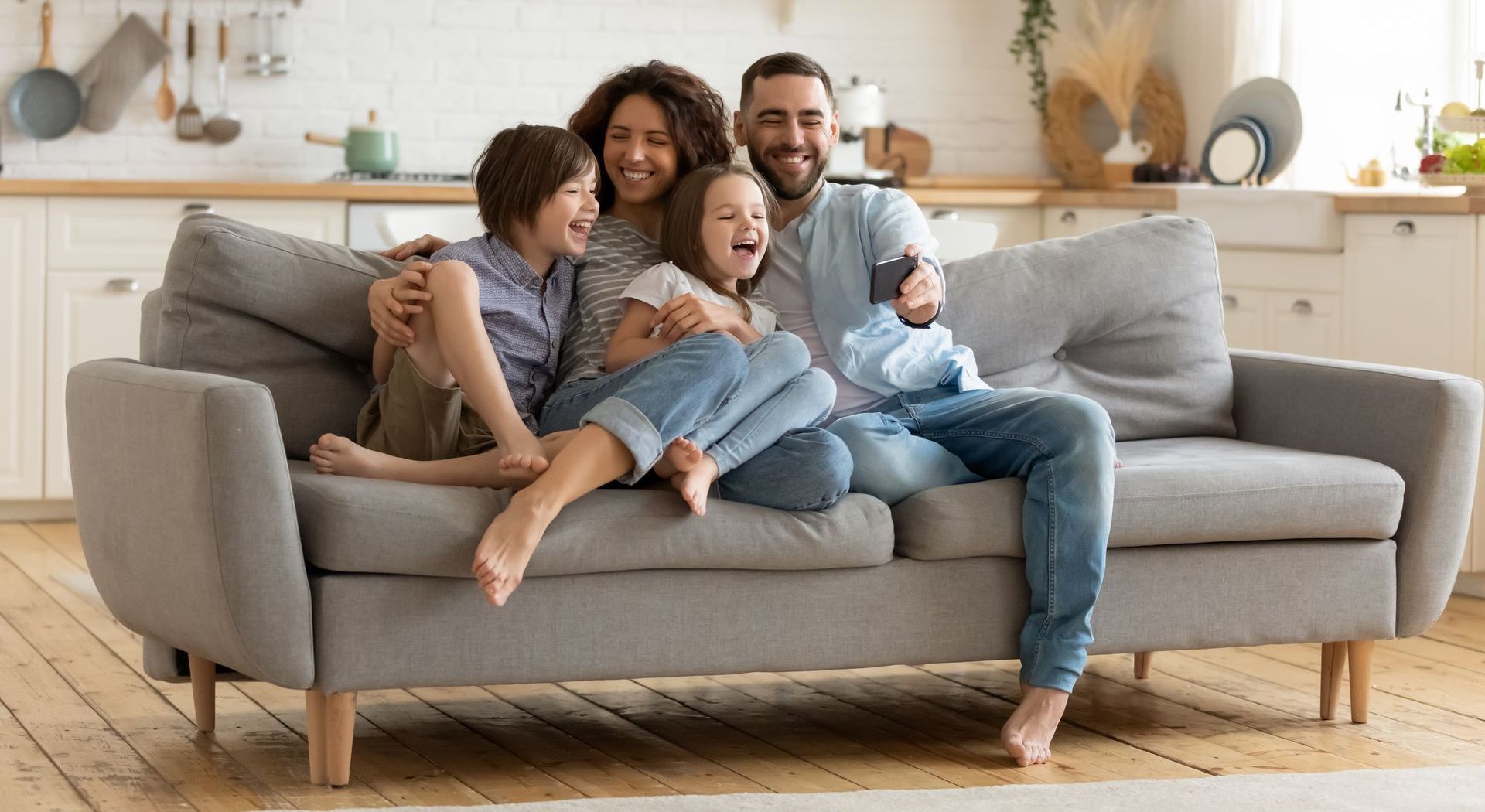 Family of four laughing on a couch in a kitchen, taking a selfie.