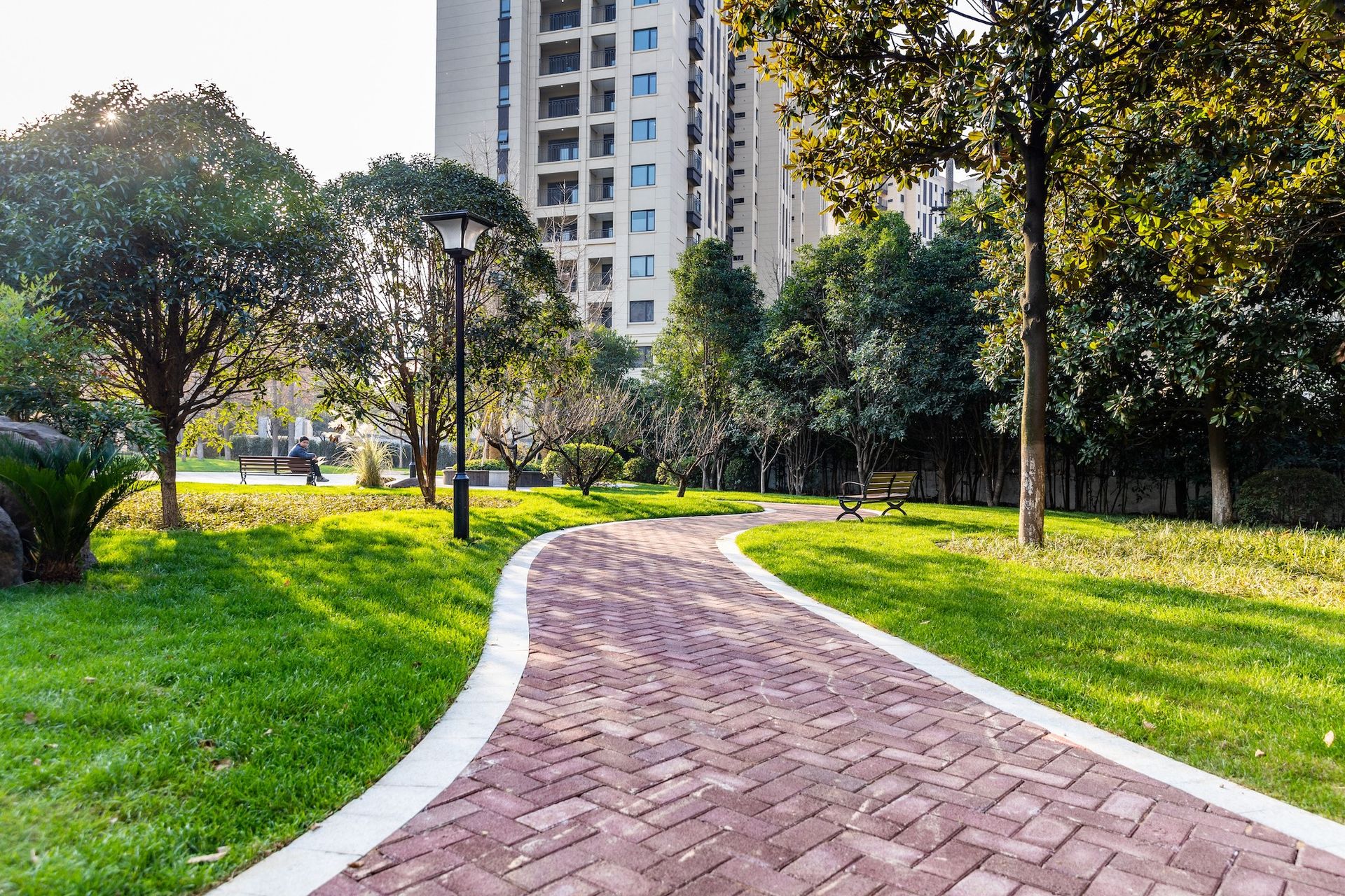 Brick path winds through a park with green grass, trees, and a tall building in the background.