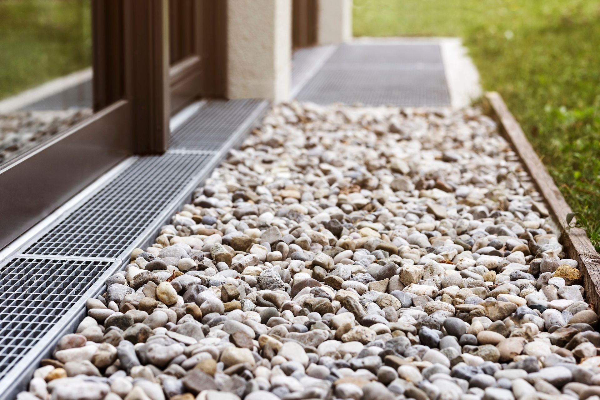 Gravel pathway alongside a building, with a metal grate drain and wooden border.