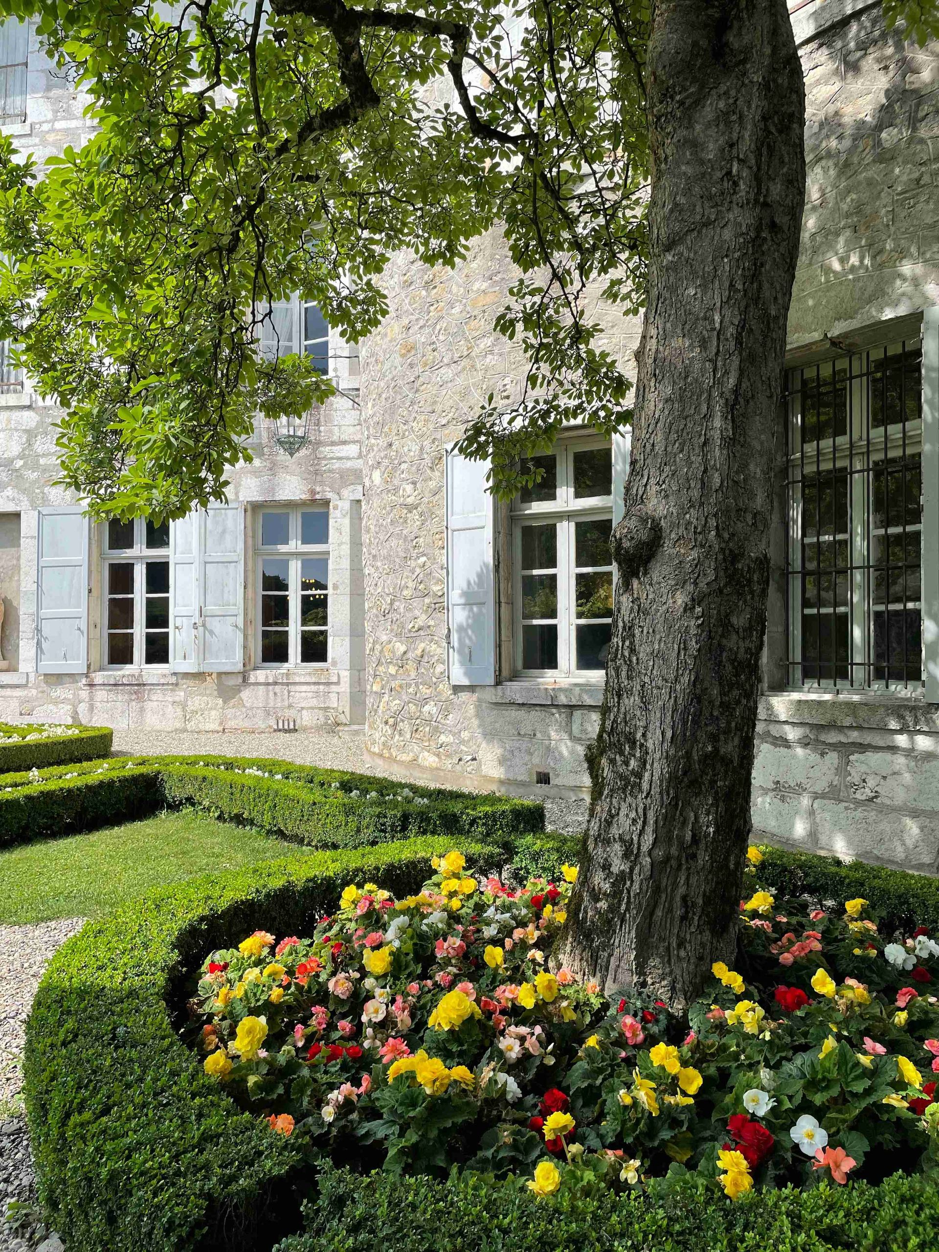 Stone building with white shutters and a colorful flower bed beneath a large tree.