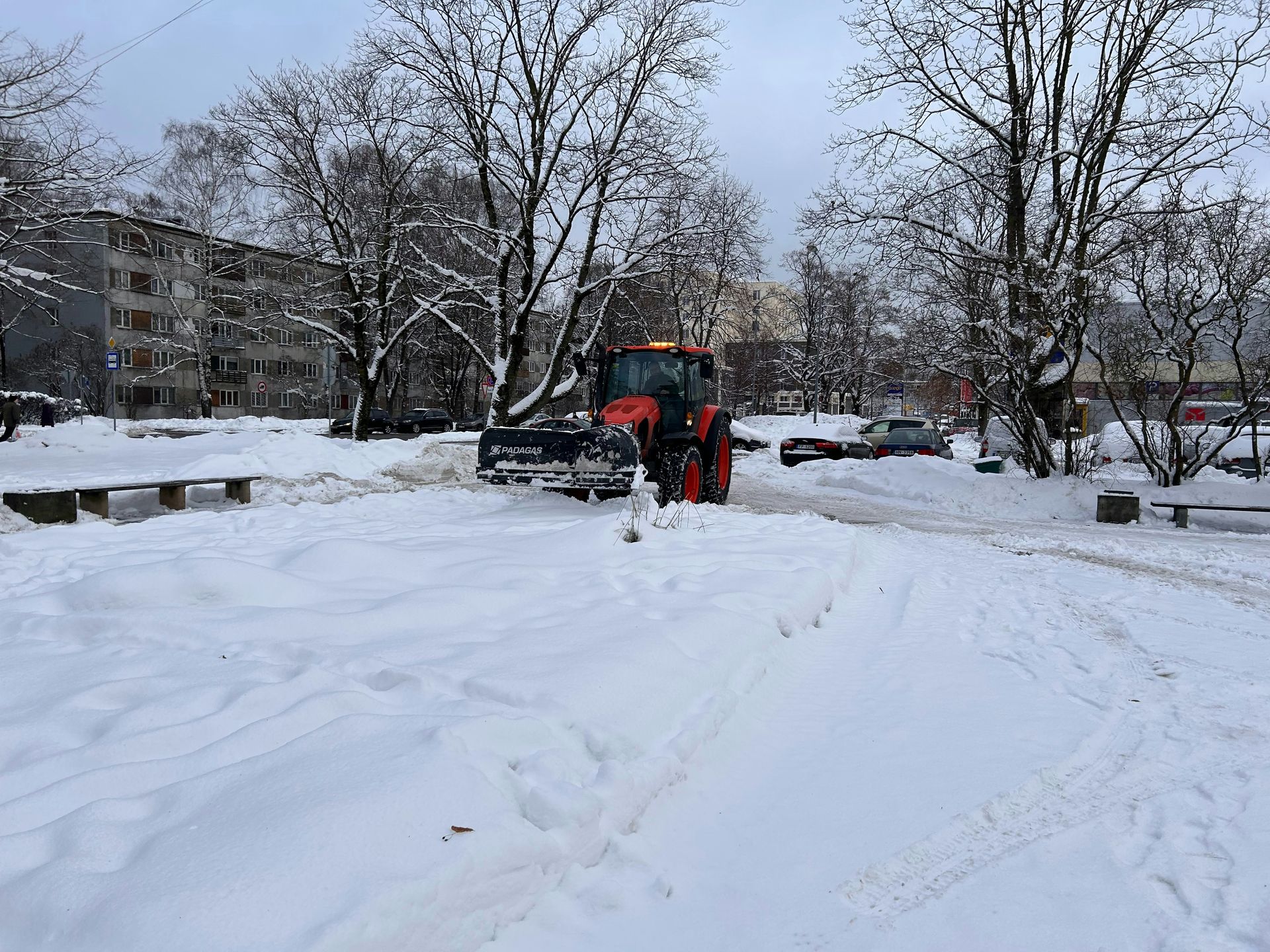 Snow-covered courtyard with a person in an orange jacket clearing a path beside parked cars and apartment buildings