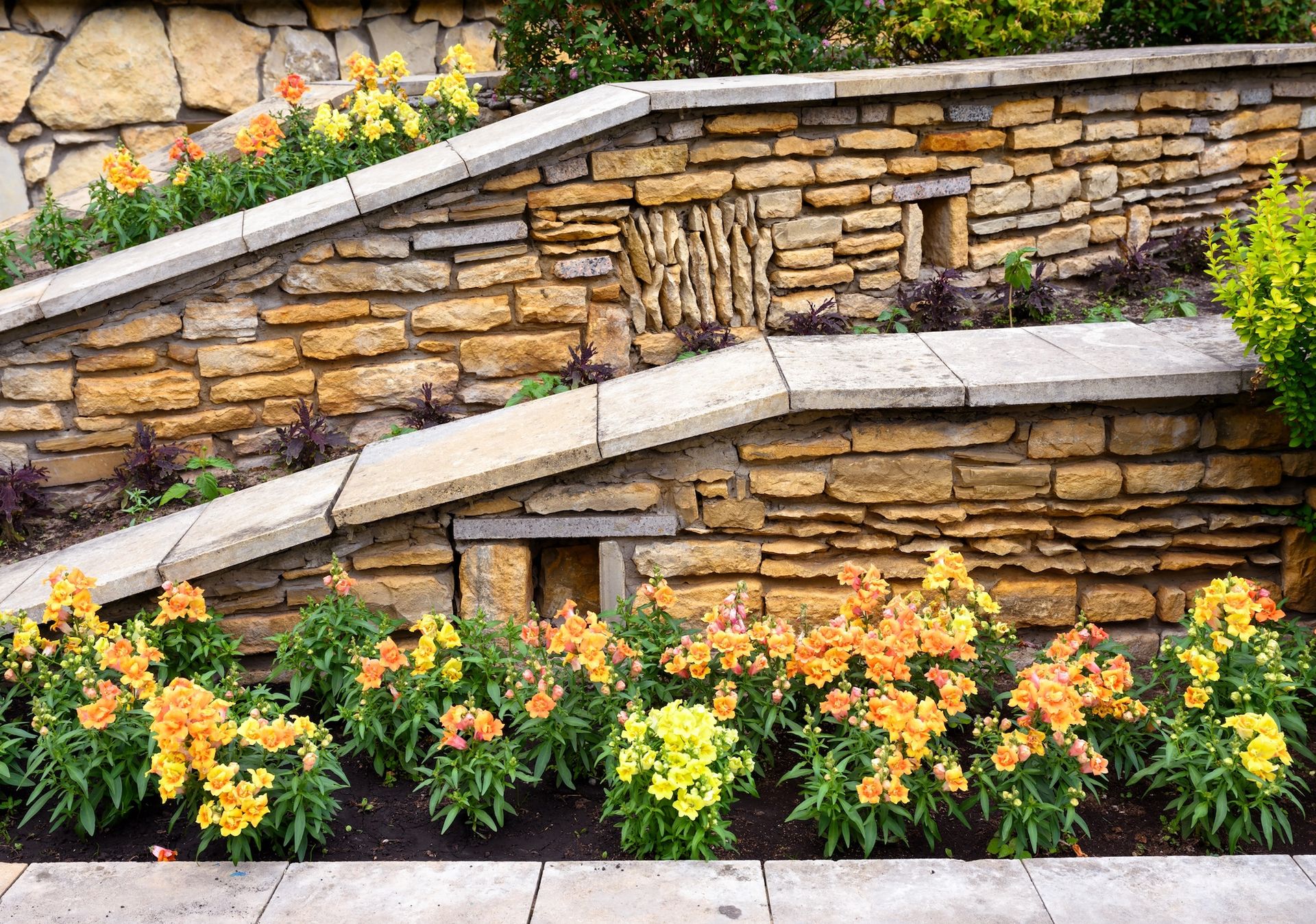 Stone retaining wall with tiered flower beds filled with yellow and orange blooms.