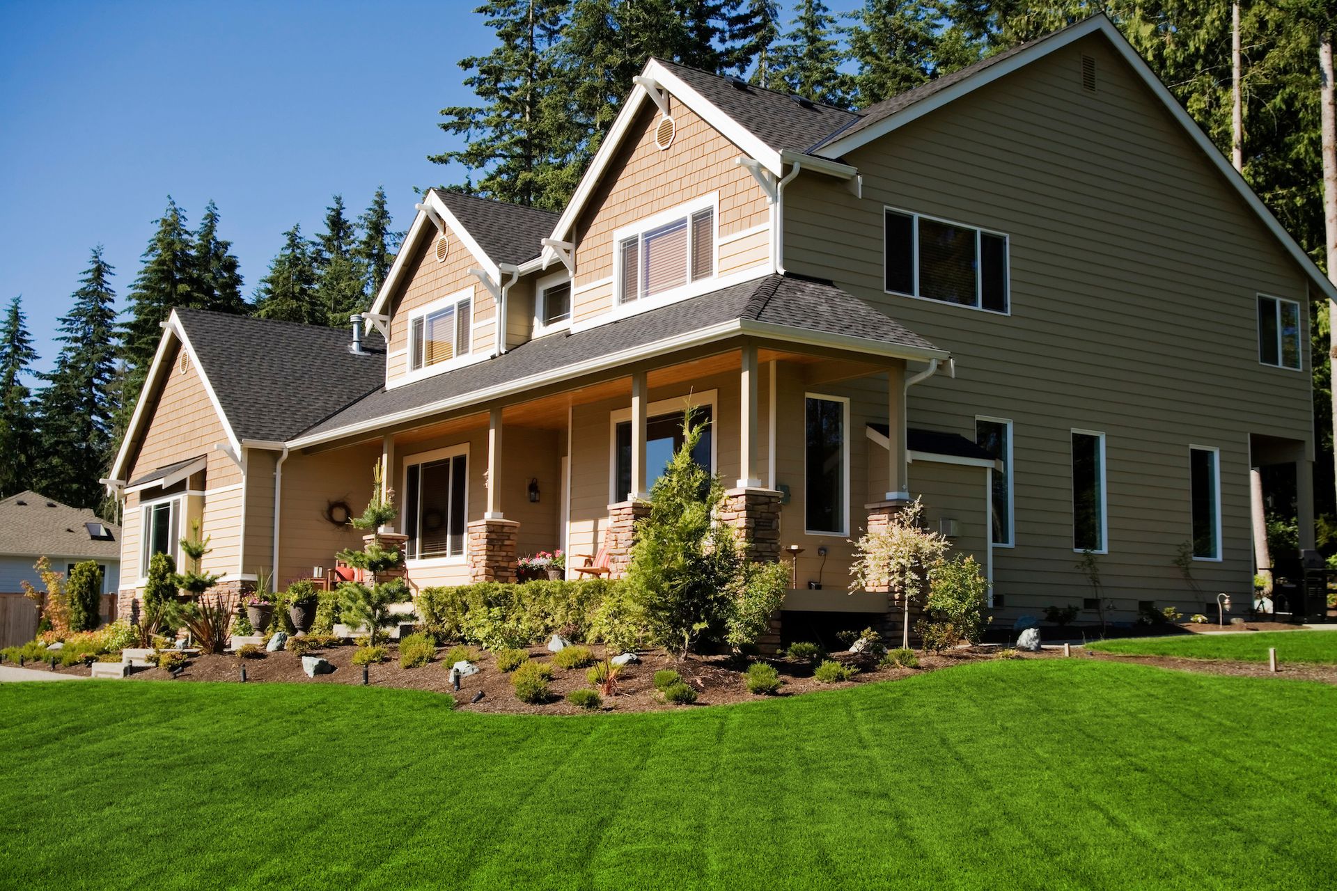 Two-story beige house with a porch and dark roof, set in a yard with green grass and trees.