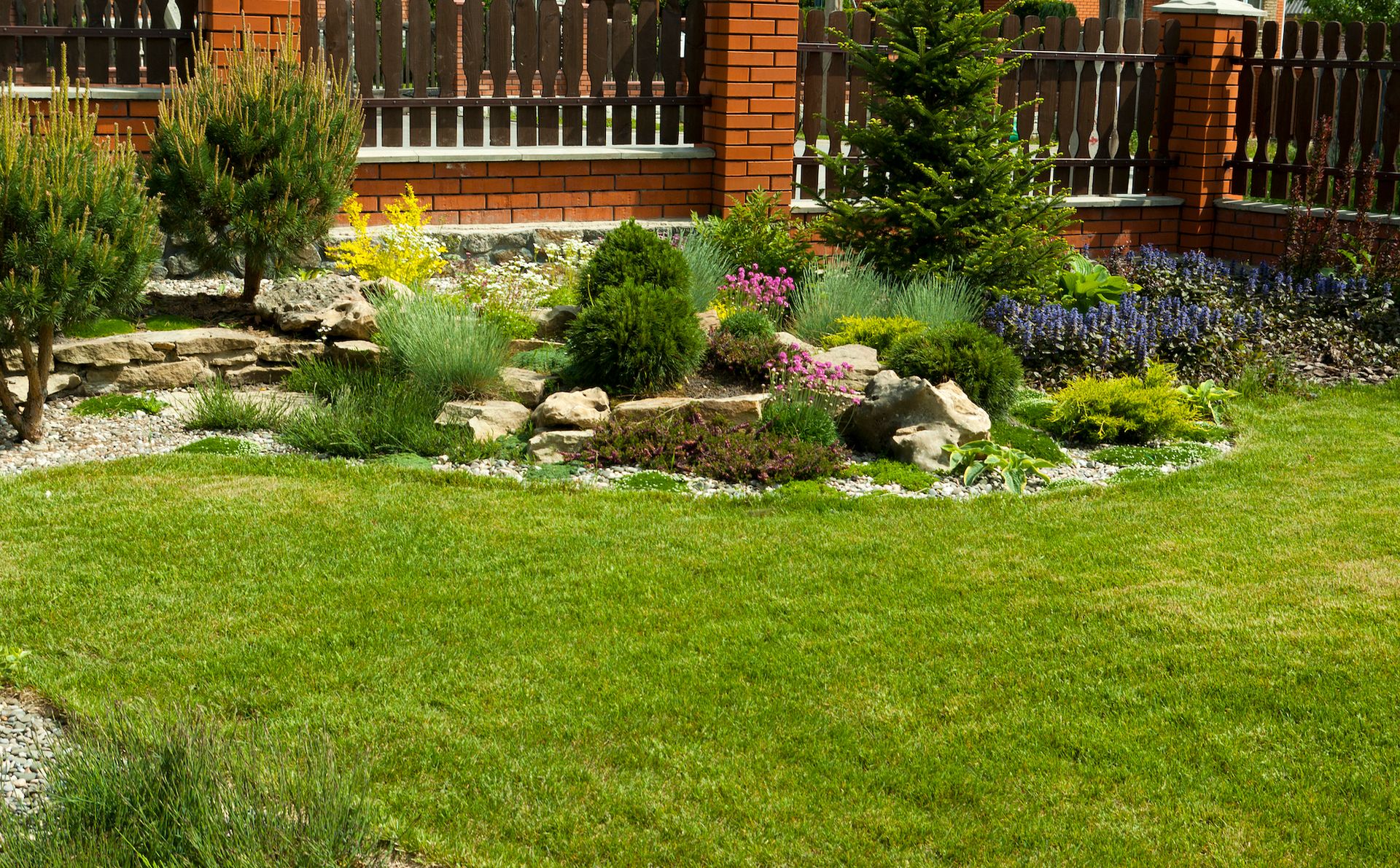 Lush green lawn with a rock garden filled with colorful flowers and shrubs in front of a wooden fence.