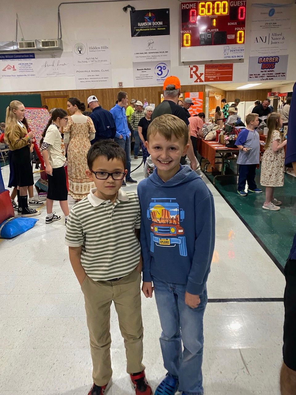 Two children standing together in a busy gymnasium, smiling at the camera.