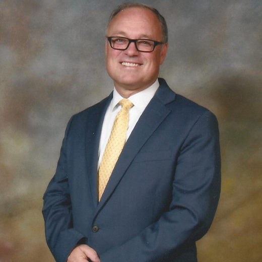 Smiling man in a navy suit and yellow tie against a blurred studio background