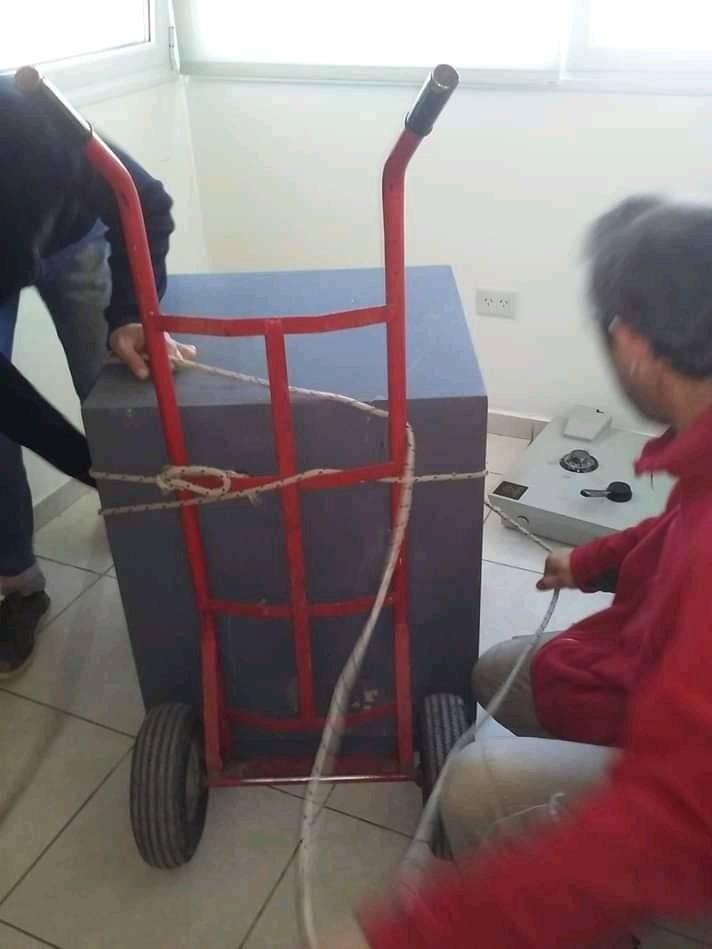 Dos personas trasladando una caja azul en una carretilla roja al interior de una vivienda.