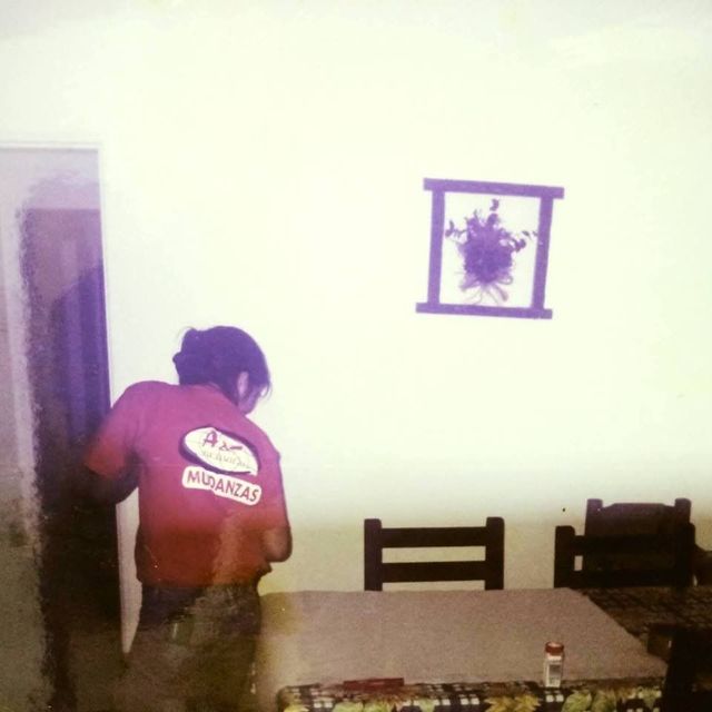 Una persona con una camisa roja está de pie cerca de una puerta, junto a una mesa, en una habitación escasamente amueblada.