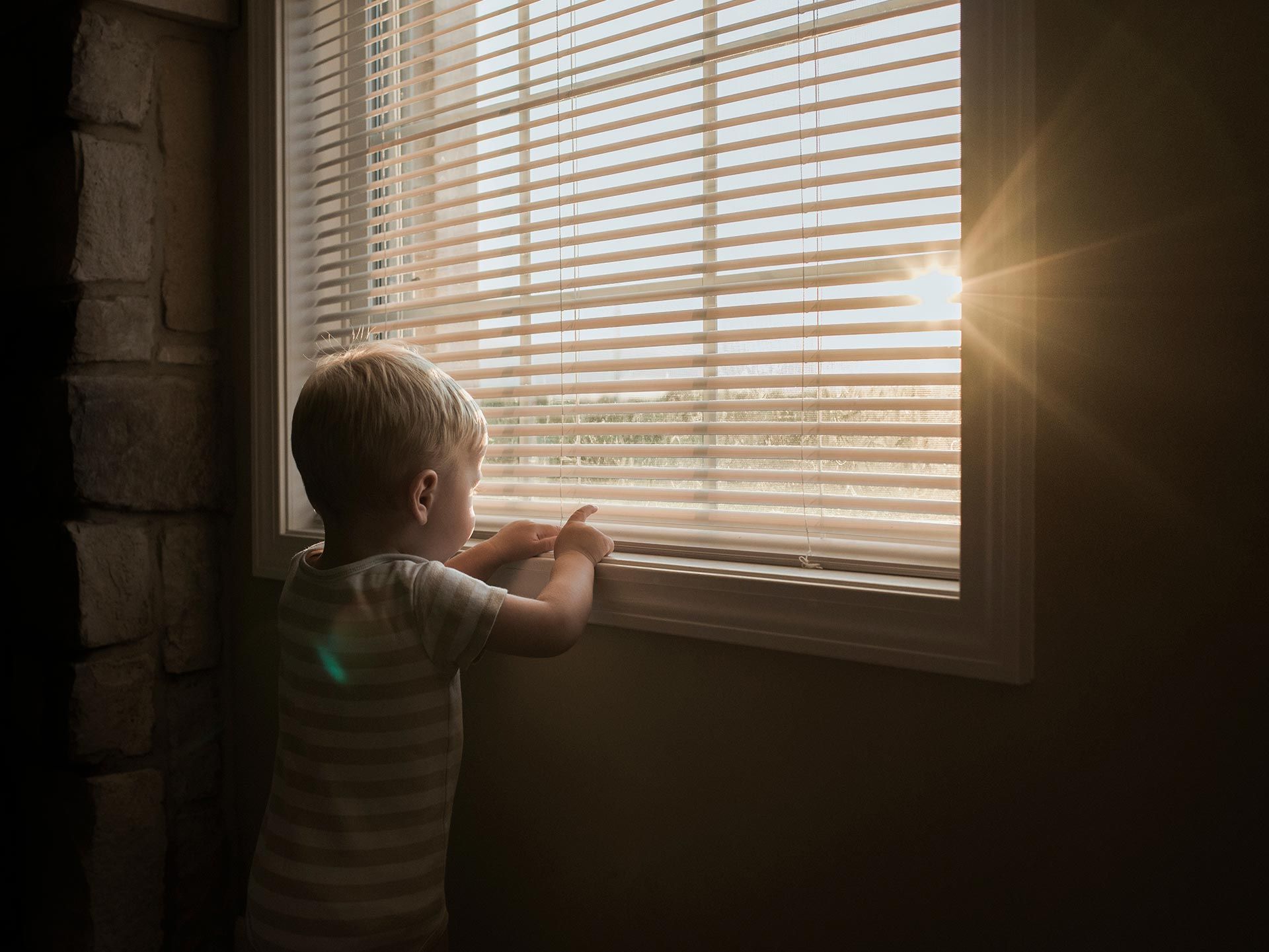 Child looking through window blinds at bright sunlight in a dim room Child looking through window blinds at bright sunlight in a dim room
