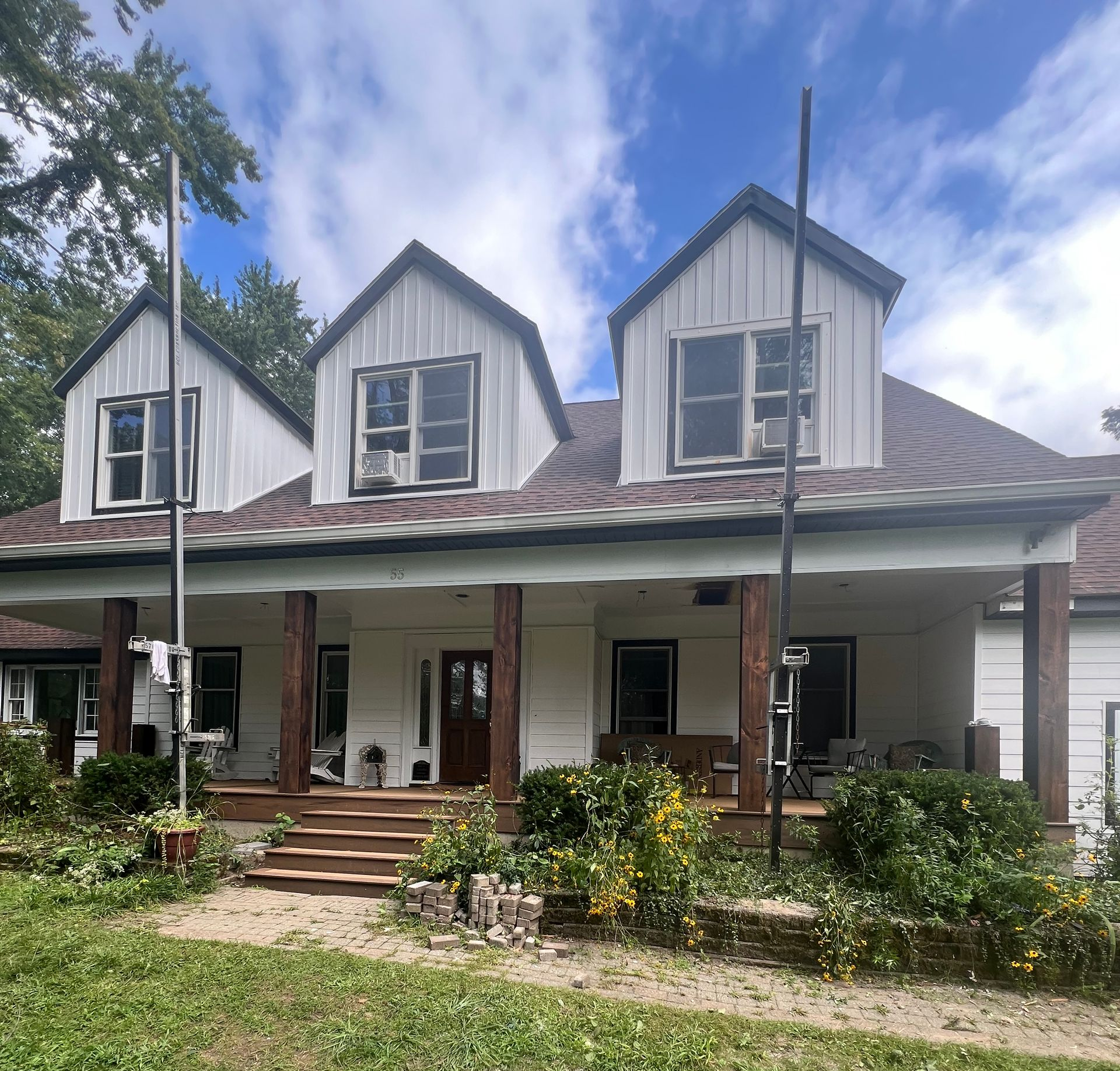 White house with a porch, three dormers, and brown roof. Blue sky, green lawn, and trees in the background.