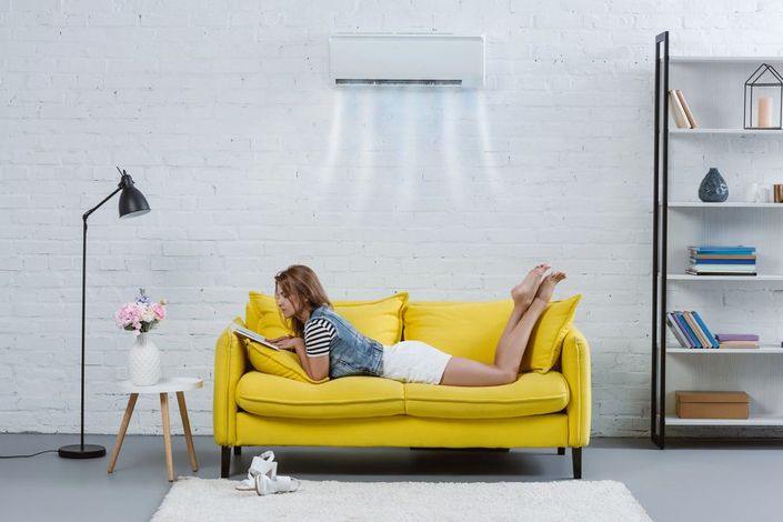 Una persona è sdraiata su un divano giallo in un luminoso soggiorno, mentre usa un computer portatile accanto a una lampada da terra e a una libreria.