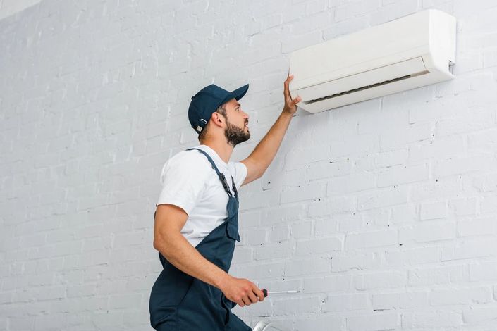 Un tecnico ispeziona un condizionatore d'aria a parete installato su un muro di mattoni bianchi.